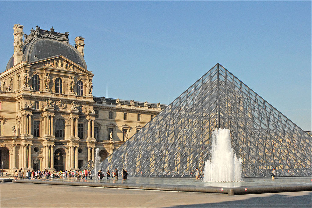 Photograph of the Pavillon Denon in the Louvre Museum plaza, with it's glass pyramid at the center - 
     	blue sky and white clouds above.