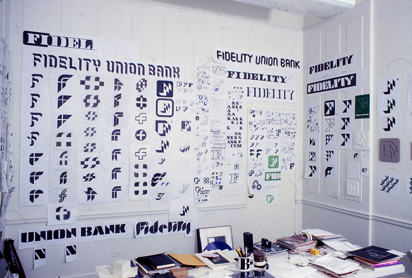 Photo of the office desk of Bill Cannan office desk at 2112 Broadway in New York City surrounded with dozens of hand drawn 
        design development drawings for the Fidelity logo and typeface illustrating the painstaking process of trial and error 
        that led to the finished products in the days before computer graphics.
