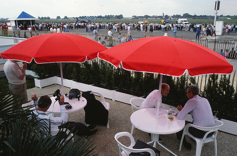 People seated at tables outside near the airstrip beneath large umbrellas.